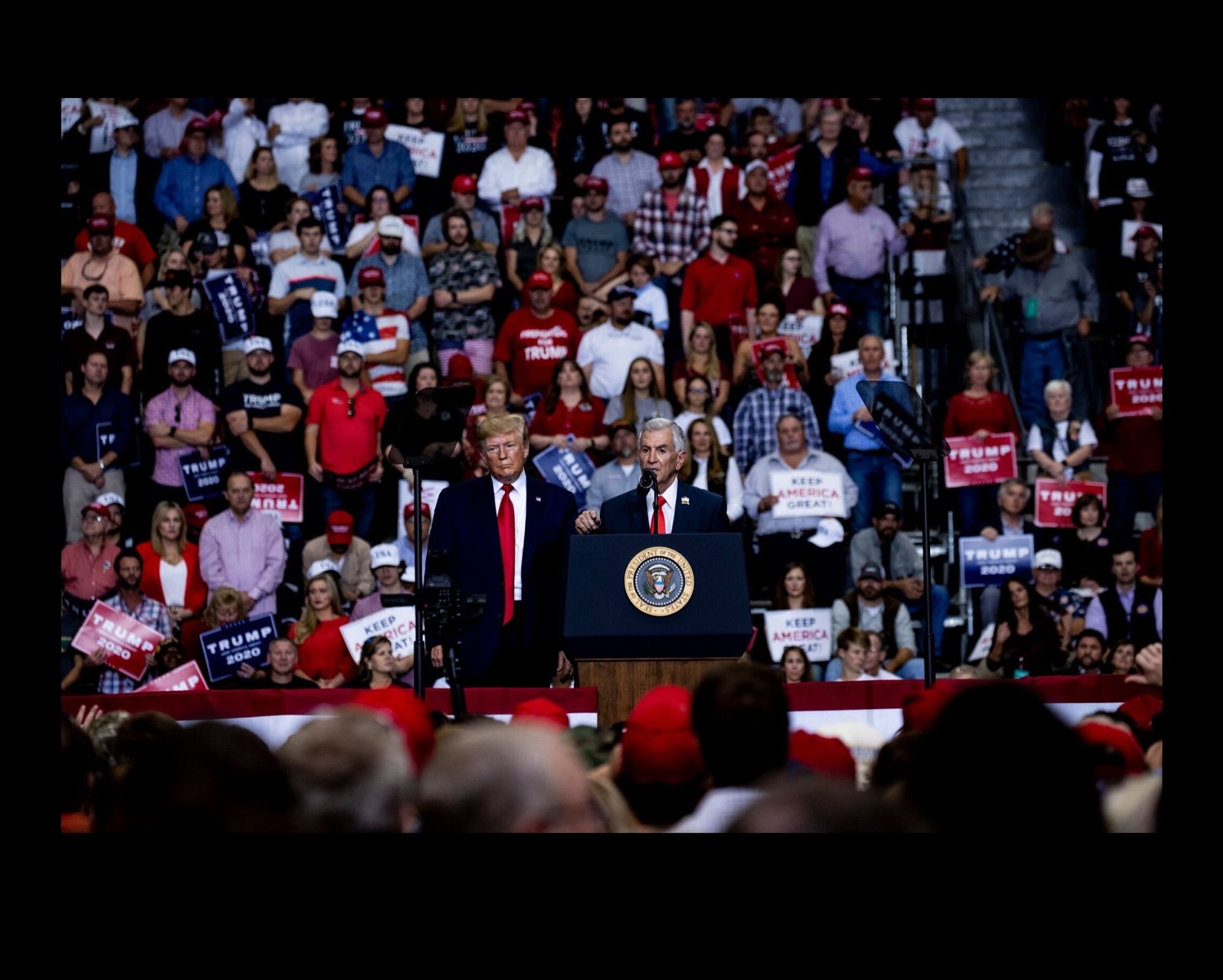 President Trump and Vice President Pence standing on a stage at a Trump rally with supporters sitting behind them on bleachers and also in the foreground.